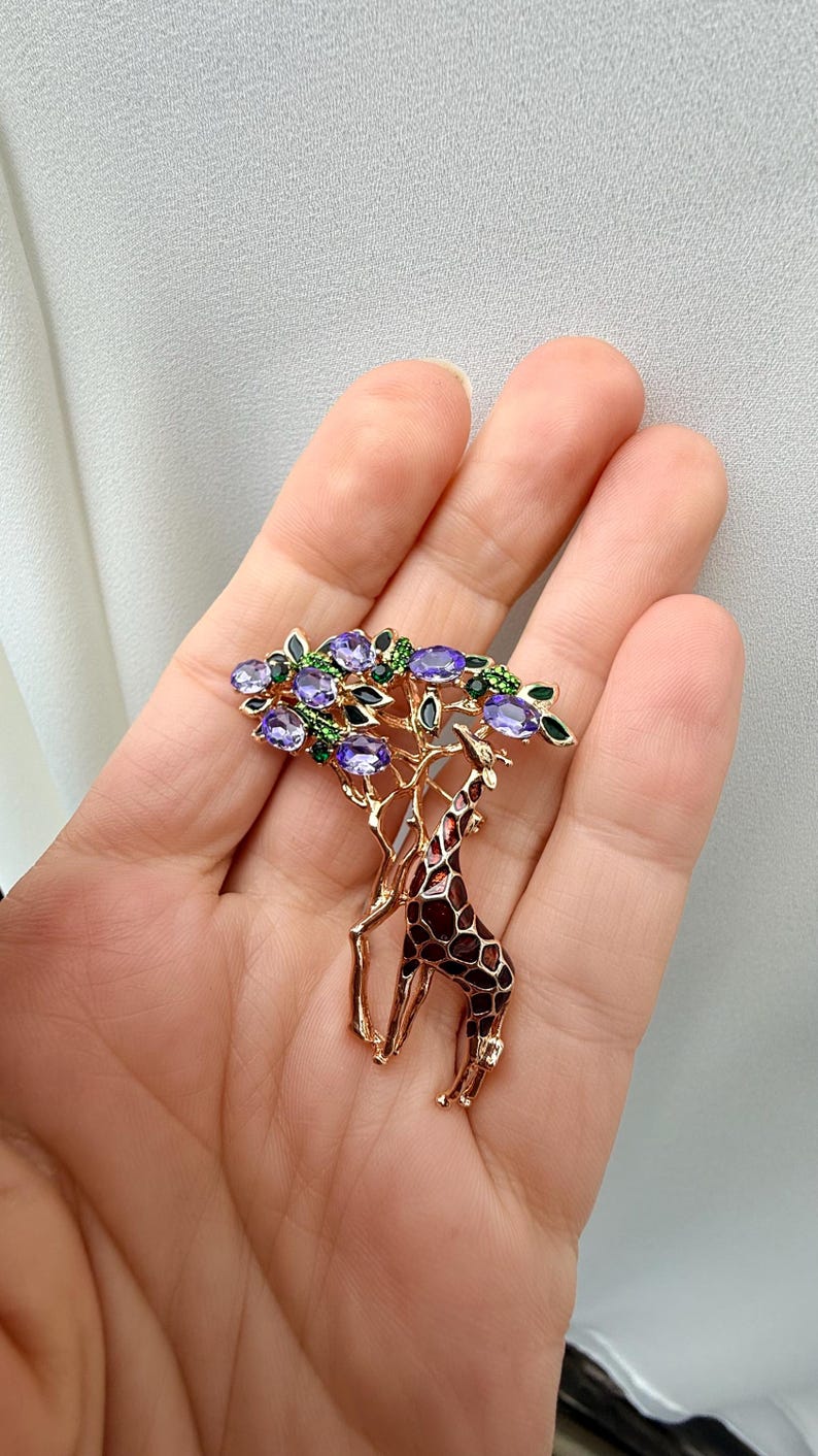 K&ouml;nnte beinhalten: Goldfarbene Brosche mit einer Giraffe unter einem Baum. Der Baum hat violette und gr&uuml;ne Edelsteinakzente. Die Giraffe hat ein braunes und schwarzes Muster. Die Brosche ist ein elegantes Accessoire.