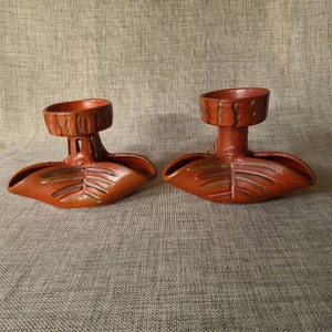 May include: Two terracotta candle holders with a leaf design. Each holder has a small, round top for a candle and a larger, open area below for a decorative element.