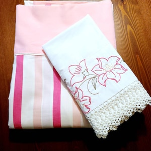 May include: A white cotton pillowcase with pink floral embroidery and a white crocheted trim. The pillowcase is folded on top of a pink and white striped sheet.