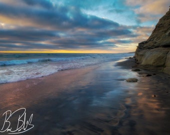 May include: A scenic view of a beach with a rocky cliff in the background. The sky is a mix of blue and gray with a hint of orange from the setting sun. The waves are crashing on the shore, and the sand is wet and reflective.