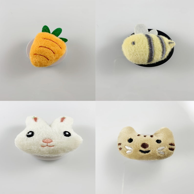 May include: Four plush toy patches with adhesive backing. The patches are shaped like a carrot, a bee, a rabbit, and a cat. The carrot is orange with green leaves. The bee is yellow with black stripes. The rabbit is white with pink ears and a pink nose. The cat is brown with white whiskers.