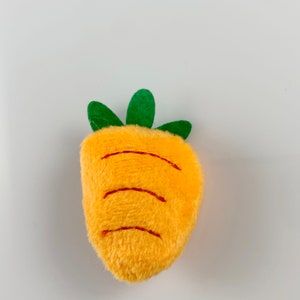 May include: A plush orange carrot toy with green leaves. The carrot has embroidered lines to resemble the ridges of a real carrot.