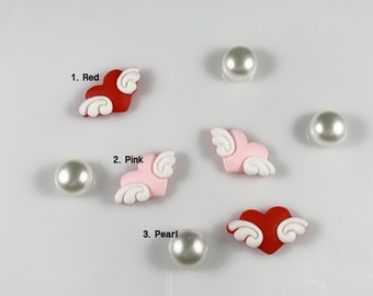 May include: Three sets of winged heart charms, one red, one pink, and one white pearl. The charms are displayed on a white surface.