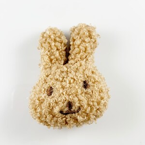 May include: A small, plush brown bunny with a smiling face. The bunny is made of fluffy, curly fabric.