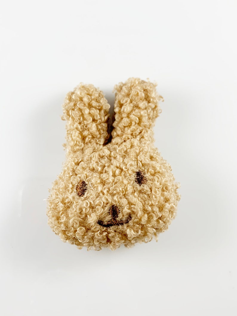 May include: A small, plush brown bunny with a smiling face. The bunny is made of fluffy, curly fabric.