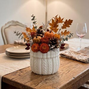 May include: A white ceramic vase with a cable knit texture filled with an autumn arrangement. The display includes small orange pumpkins, brown pinecones, dried orange flowers, red berries, eucalyptus sprigs, and dried maple leaves.
