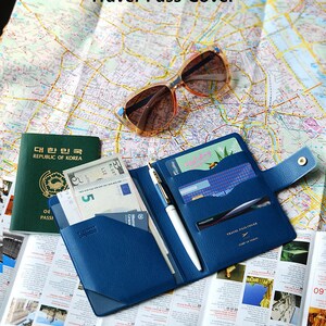 May include: A navy blue leather travel passport holder with a pen, cash, and credit cards inside. The holder has a flap with a button closure and the text "TRAVEL PASS COVER" and "START OF TRAVEL" printed on it.