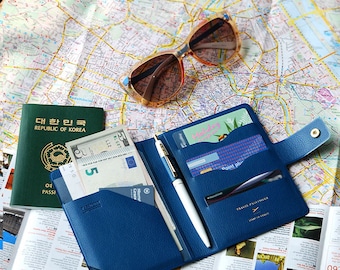 May include: A navy blue leather travel passport holder with a pen, cash, and credit cards inside. The holder has a flap with a button closure and the text "TRAVEL PASS COVER" and "START OF TRAVEL" printed on it.