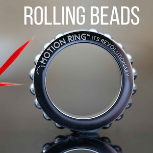 Puede incluir: Un anillo plateado con una banda negra y las palabras "Motion Ring&trade; Its Revolutionary" grabadas alrededor de la banda. El anillo est&aacute; hecho de cuentas rodantes.
