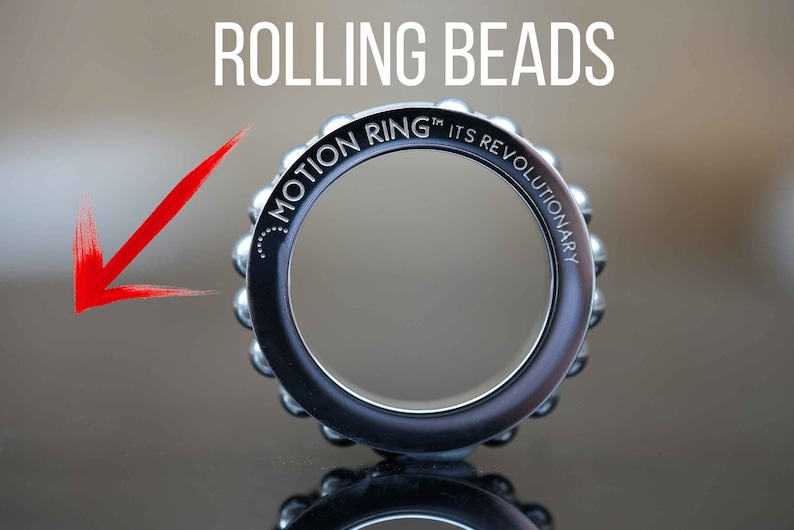 Puede incluir: Un anillo plateado con una banda negra y las palabras "Motion Ring&trade; Its Revolutionary" grabadas alrededor de la banda. El anillo est&aacute; hecho de cuentas rodantes.