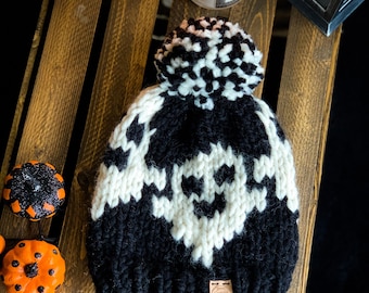 May include: Black and white knit beanie with a skull pattern and a white pom pom. The beanie has a brown leather tag with the text "Georgia Moore Designs" on it.