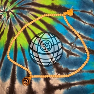 May include: A necklace featuring a long strand of small, light brown wooden beads arranged in a circular loop. The necklace includes a small yellow tassel and a dark, circular pendant with a spiral design made of copper-colored wire.