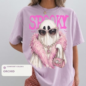 A person wearing a purple t-shirt with a graphic design of a ghost wearing sunglasses, a pink feathered scarf, and holding a pink purse.