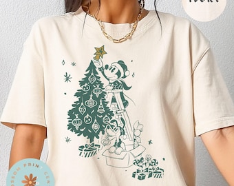 Puede incluir: Camiseta color marfil con un gráfico verde de Mickey Mouse decorando un árbol de Navidad. Pluto está en una caja con regalos. El texto "Blossom Print Center" está en la parte inferior. La palabra "IVORY" está arriba.