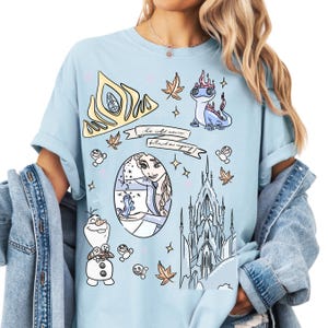 A woman wearing a light blue t-shirt with various cartoon-style illustrations and text, including characters from the Disney movie Frozen.