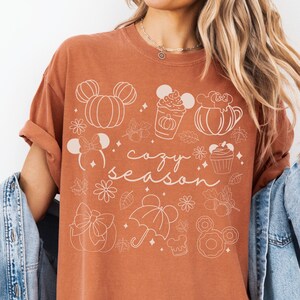 A woman wearing a t-shirt with a graphic design featuring various Disney-themed elements such as Mickey Mouse ears, pumpkins, and umbrellas, along with the text "cozy season".
