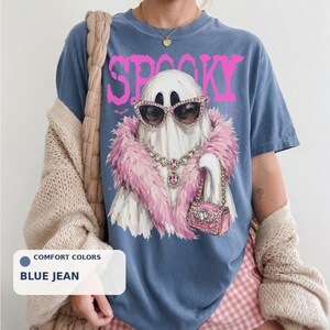 A person wearing a blue t-shirt with a graphic of a ghost wearing sunglasses and a pink feather boa, along with the word "SPOKY" printed on it.