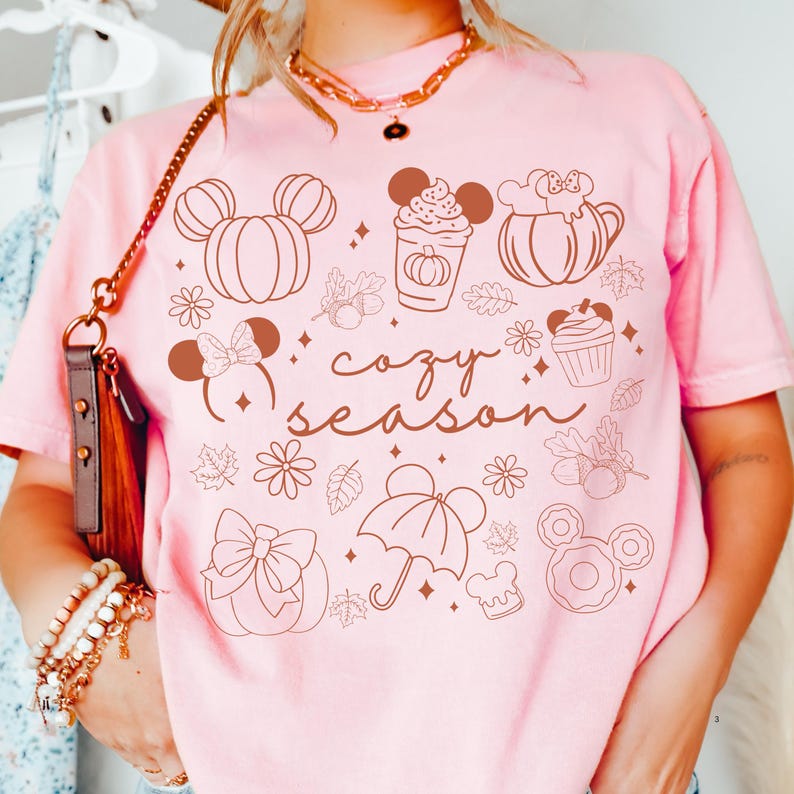 A person wearing a pink t-shirt with various Disney-themed illustrations and text, including Mickey Mouse, pumpkins, cupcakes, and the word "cozy" written on it.