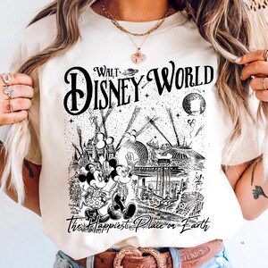 A person wearing a white t-shirt with a graphic design depicting Mickey Mouse and Minnie Mouse in a whimsical, fantastical landscape.