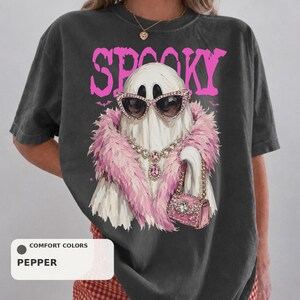 A person wearing a black t-shirt with a graphic design of a ghost wearing sunglasses, a pink feathered scarf, and holding a pink purse.