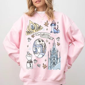 A woman wearing a pink sweatshirt with various cartoon-style illustrations and text, including characters and scenes from the Disney movie Frozen.