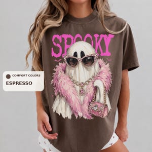 A woman wearing a brown t-shirt with a graphic design of a ghost wearing sunglasses and a pink fur coat.