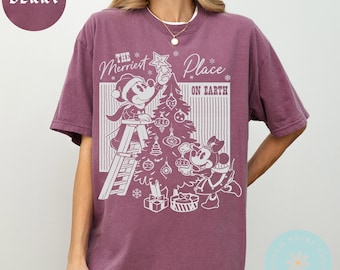 Puede incluir: Camiseta de color bayas con un gráfico de Mickey y Minnie Mouse decorando un árbol de Navidad. El texto de la camiseta dice "The Merriest Place on Earth". La camiseta tiene cuello redondo y mangas cortas.