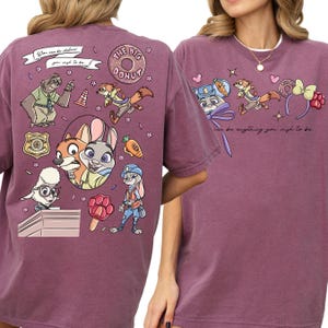 a purple t-shirt with a vibrant graphic design of cartoon characters, including a bear, a rabbit, and other animals, along with various objects like a carrot, a flower, and a sign.