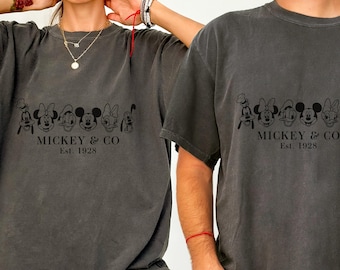 Könnte beinhalten: Zwei anthrazitfarbene T-Shirts mit einer schwarzen Grafik von Micky Maus und Freunden. Der Text "Mickey & Co. Est. 1928" ist unter den Figuren aufgedruckt. Die Shirts haben kurze Ärmel und einen Rundhalsausschnitt.