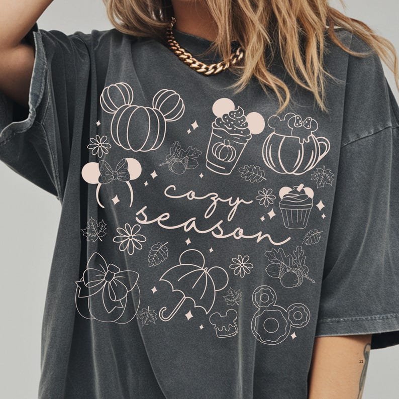 A person wearing a gray t-shirt with a graphic design featuring various Disney-themed elements such as Mickey Mouse, pumpkins, and umbrellas, along with the text "cozy season".
