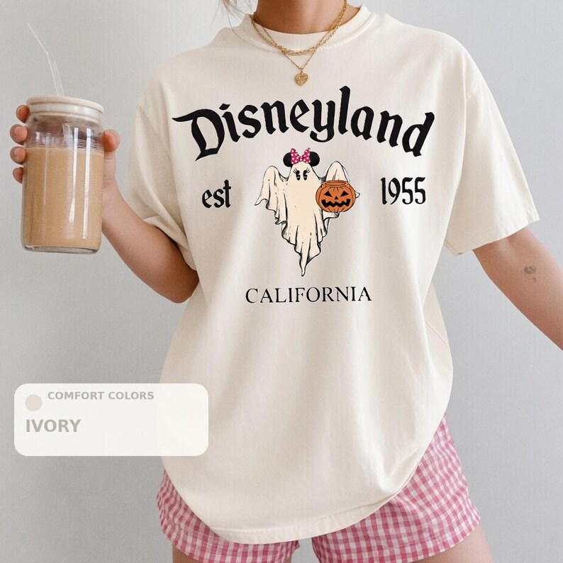 A person wearing a white t-shirt with a graphic of a ghost and a pumpkin, along with the text "Disneyland est 1955" and "California". They are holding a drink in a glass.