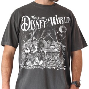 A man wearing a gray t-shirt with a graphic design depicting Mickey Mouse and other Disney characters in a nighttime scene with fireworks and a full moon.