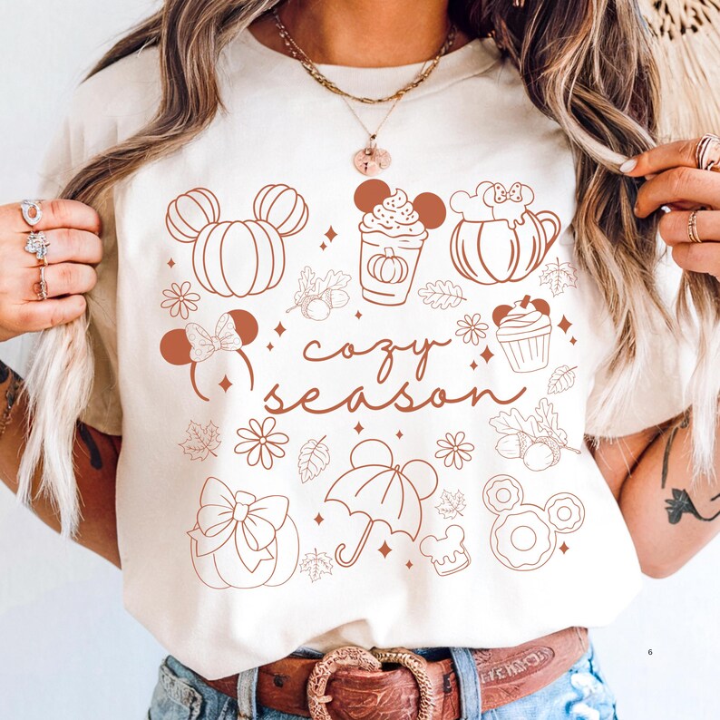 A woman wearing a white t-shirt with a graphic design featuring various Disney-themed items such as Mickey Mouse, pumpkins, and umbrellas, along with the text "cozy season".