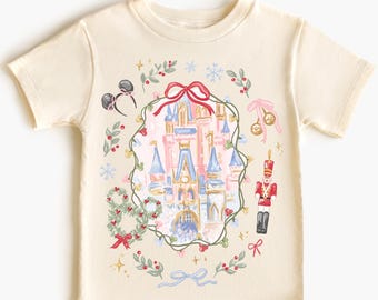 May include: Cream-colored t-shirt featuring a whimsical illustration of a fairytale castle, surrounded by a festive wreath with red bows, ornaments, and a toy soldier. The design includes Mickey Mouse ears and snowflakes, evoking a holiday theme.
