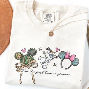 A white t-shirt with a graphic design featuring characters from Disney's Frozen, including Olaf the snowman, Minnie Mouse, and other iconic figures. The text on the t-shirt reads "For the first time in forever".