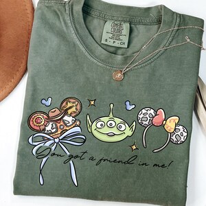 A green t-shirt with a graphic design featuring a green alien character, a bow, and various Disney-themed elements.
