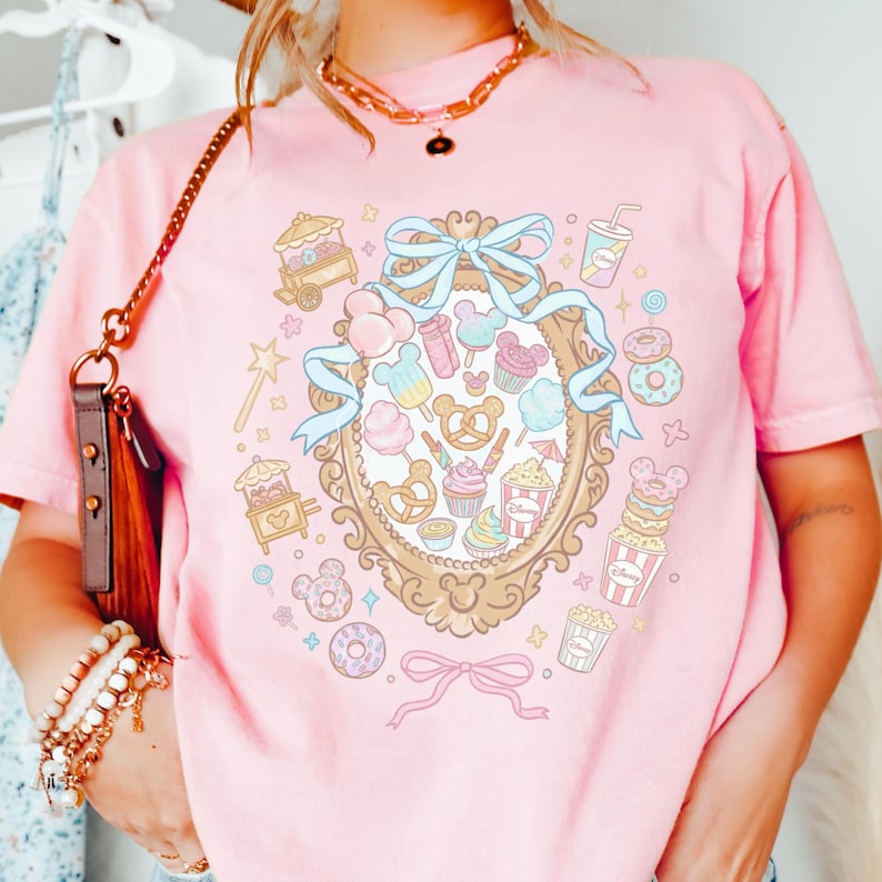 A person wearing a pink t-shirt with a whimsical graphic design featuring various Disney-themed items such as Mickey Mouse, popcorn, and candy.