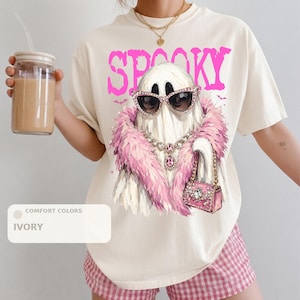 A person wearing a white t-shirt with a graphic of a ghost wearing sunglasses, a pink feathered scarf, and holding a drink.