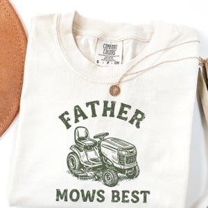 May include: Cream-colored t-shirt with the text "FATHER MOWS BEST" in green, along with a graphic of a riding lawn mower. The shirt has a crew neck and short sleeves. A gold necklace with a pendant is draped across the shirt.