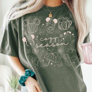 A woman wearing a green t-shirt with a graphic design of various Disney-themed items, including Mickey Mouse, pumpkins, and umbrellas, along with the text "cozy season".
