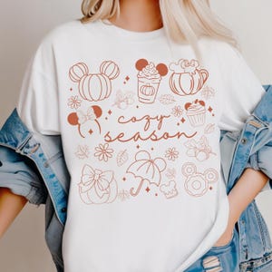 A white t-shirt with a graphic design featuring various fall-themed elements such as Mickey Mouse ears, pumpkins, cupcakes, and umbrellas, along with the text "cozy season".