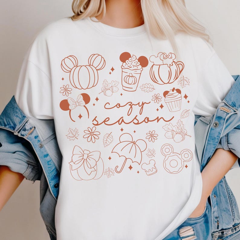 A white t-shirt with a graphic design featuring various fall-themed elements such as Mickey Mouse ears, pumpkins, cupcakes, and umbrellas, along with the text "cozy season".