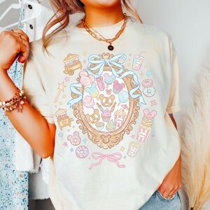 A woman wearing a white t-shirt with a colorful graphic design featuring various cartoon-like objects and characters, such as donuts, cupcakes, and Mickey Mouse.
