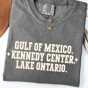 May include: A charcoal gray t-shirt featuring the cream-colored text GULF OF MEXICO. KENNEDY CENTER. LAKE ONTARIO. with diamond accents. A thin silver-toned chain necklace with a small circular pendant rests on the neckline of the shirt.
