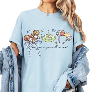 A woman wearing a light blue t-shirt with a graphic design featuring a green alien character and various Disney-themed elements.