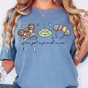 A woman wearing a blue t-shirt with a graphic design featuring a green alien character, a pair of colorful earrings, and a ribbon.