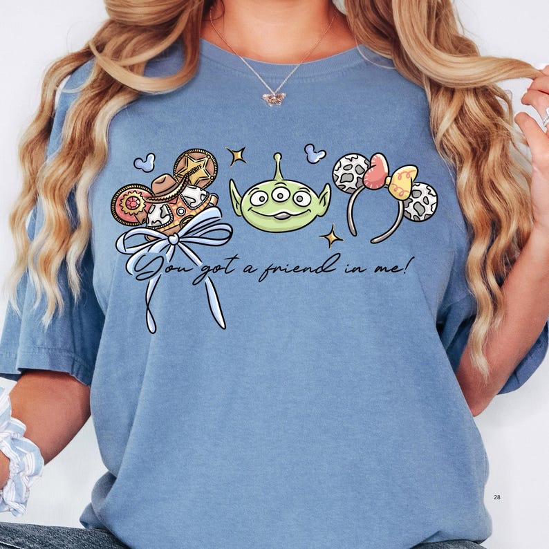 A woman wearing a blue t-shirt with a graphic design featuring a green alien character, a pair of colorful earrings, and a ribbon.