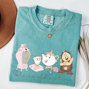 May include: Teal Comfort Colors t-shirt featuring a graphic of characters from a fairy tale. The design includes a pink wardrobe, a teacup, a teapot, a clock, a book, and a rose. The text "Tale as old as time" is written below the characters.