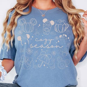 A woman wearing a blue t-shirt with a graphic design featuring various Disney-themed illustrations, including Mickey Mouse, pumpkins, and umbrellas, along with the text "cozy season".