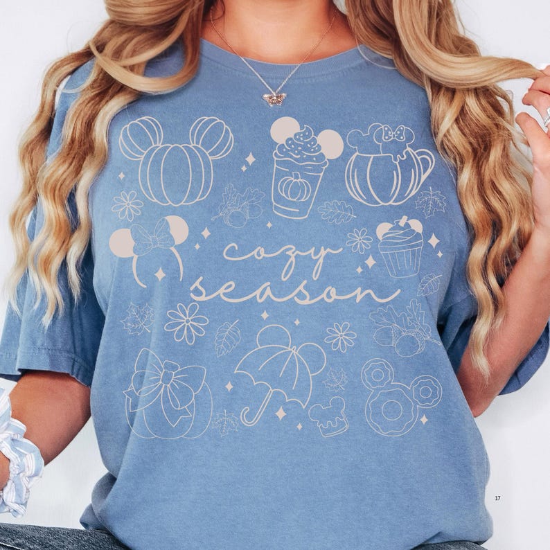 A woman wearing a blue t-shirt with a graphic design featuring various Disney-themed illustrations, including Mickey Mouse, pumpkins, and umbrellas, along with the text "cozy season".
